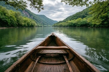 Obraz premium Wooden canoe points towards a serene river surrounded by lush, green mountains under a cloudy sky, framed by leafy branches