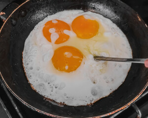 Iron frying pan with three eggs, closeup photo. Three eggs frying in pan, cooking breakfast on stove with metal spoon