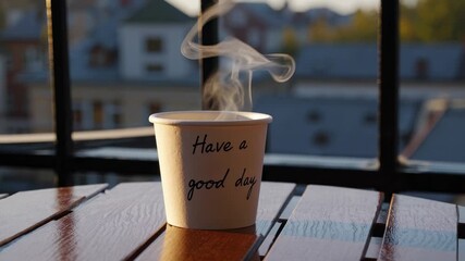 Steam rises in morning light. Paper coffee cup with positive quote stands on wood table. Sunlight shines on cup. Hot drink takeaway moment. Cup, quote, paper, drink, hot, steam, morning.