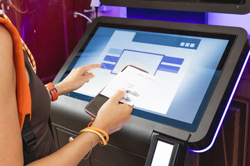 Woman scanning barcode on self service kiosk with touch screen, using smartphone and printed receipt
