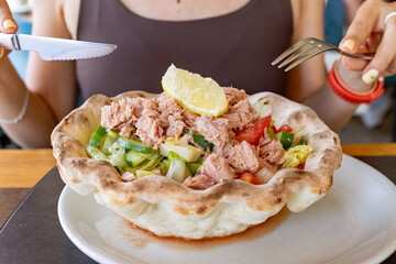 Woman holding fork and knife, preparing to eat a fresh tuna salad served inside a bread bowl, with lettuce, tomatoes, cucumber and lemon