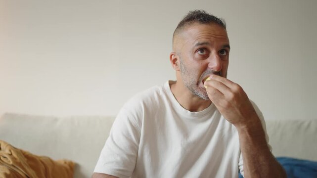 A middle aged man sits facing the camera, eating fruit while watching television, capturing the mood of ordinary and monotonous daily life