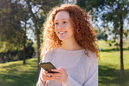young woman with curly red hair is smiling while using cell phone, she is standing in a park with trees in the background, concept of technology of communication