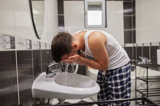 A man leans over a sink, washing his face with water in a stylish bathroom. He wears a tank top and plaid pajama pants, starting his morning routine.