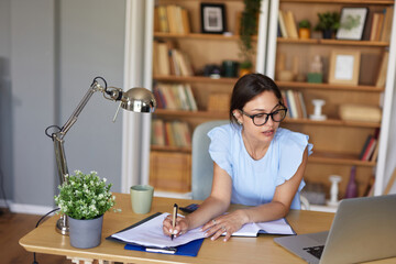 A professional woman is engaged in work at her home office. She is taking notes while using a laptop, surrounded by books and greenery in a bright, inviting space.