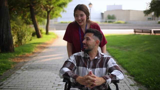 Cheerful female nurse pushing a happy disabled man in a wheelchair along a park path. The couple is laughing and enjoying their time together during his recovery and rehabilitation outdoors