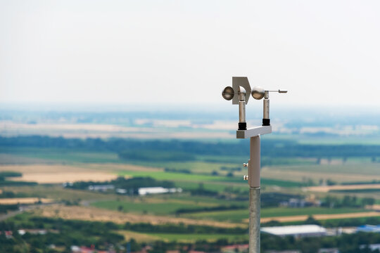 Close-up of an anemometer used for measuring wind speed, positioned on a hilltop with blurred fields and sky in the background