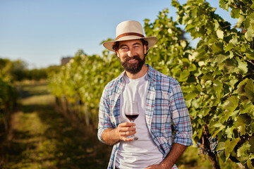 A vineyard worker stands amidst rows of green vines, smiling and holding a glass of red wine on a sunny day. The scene highlights the joy and passion of family-run wine production.