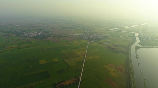 Aerial drone shot wide panning shot showcasing vibrant green fields, suburban farms, and a water body glowing under monsoon sunrise near Delhi, India.