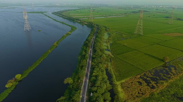 Aerial drone shot wide panning shot of fertile green farmland and reflective water body with power cables above, taken during sunrise on the outskirts of Delhi, India.