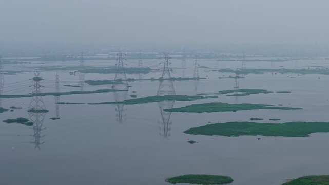Panning aerial shot showcasing reflective water body and power lines crossing above, golden sunrise glow and monsoon atmosphere near Delhi, India.