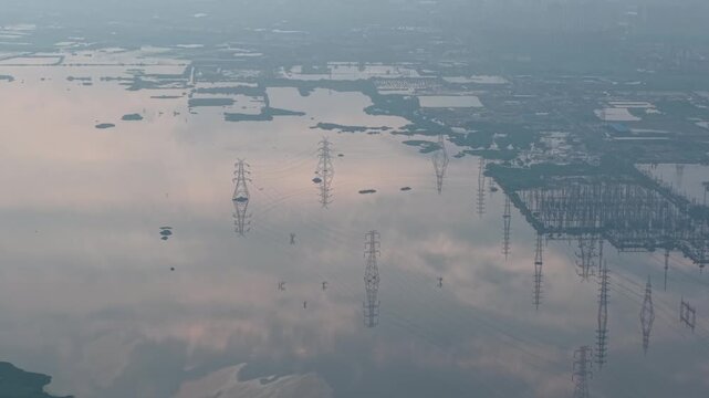Panning aerial shot of tranquil water body with power cables mirrored in the surface, surrounded by rural landscape at sunrise in the monsoon season near Delhi, India.