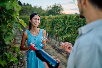 Young woman smiling, holding wine glasses, while man prepares to open a bottle of red wine in a scenic vineyard