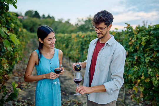 Smiling young couple tasting red wine, pouring drink in glass, enjoying romantic leisure time in a green vineyard - Powered by Adobe
