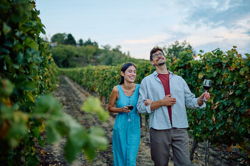 Couple walking through a vineyard, holding glasses of red wine and smiling. Enjoying a relaxing wine tasting experience