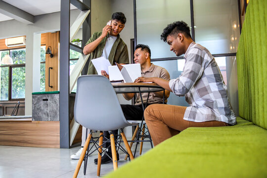 Three Young Male Colleagues Working Together on Project in Modern Office, With One Man on Phone All