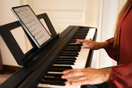 Female hands on piano keyboard with sheet music on tablet on stand, practicing musical notes on digital instrument at home indoors during daytime, close-up
 - Powered by Adobe