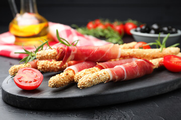 Delicious grissini sticks with sesame seeds, prosciutto and tomato on dark textured table against black background, closeup