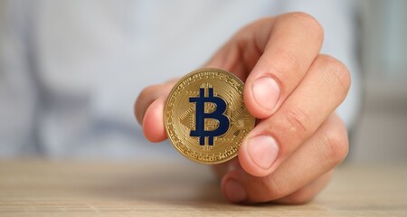 Cryptocurrency. Man with golden bitcoin at wooden table against blurred background, closeup. Space for text