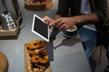African American man wearing jacket tapping white tablet at cafe counter with pretzels cappuccino