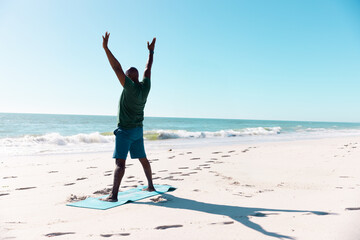 Senior African American man stretching on teal yoga mat on beach with footprints facing sea