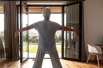 Senior woman stretching on wooden floor inside living area facing open glass doors to sunlit garden