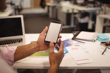 Smartphone being held above white desk in office workspace, with dark laptop and scattered charts