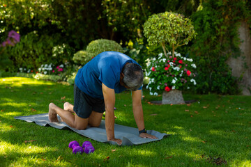 Senior man kneeling on gray exercise mat on green lawn in lush garden beside purple dumbbells