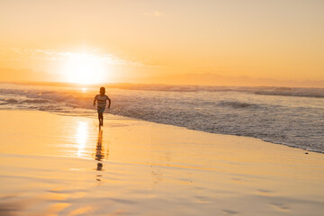 Naklejka premium African American boy running at sunset on wet beach sand, waves lapping and sun reflecting
