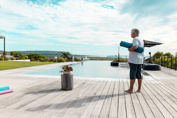 Senior man standing barefoot on wooden deck beside infinity pool holding teal yoga mat, copy space