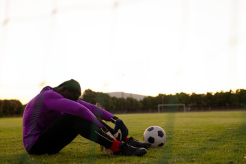 African American male goalkeeper sitting hugging knees on pitch with ball near goalpost, copy space