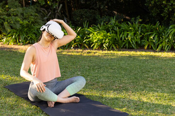 Mid adult Asian woman sitting on yoga mat in backyard wearing VR headset and stretching neck