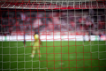 ed and white football goal net in sharp focus with a blurred goalkeeper and stadium crowd in the background. Selective focus, shallow depth of field, game atmosphere, copy space.