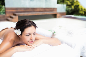 Woman lying face down on white towel-covered spa bed outdoors, resting head on arms with flower