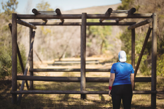 Fototapeta Female athlete standing facing wooden obstacle course frame on grassy dirt path under clear skies
