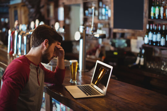 Bearded man resting on hand while gazing at laptop beside beer glass at pub, copy space