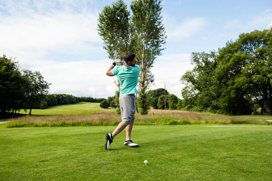 Male golfer wearing turquoise polo swinging driver at tee box with golf ball under cloudy sky