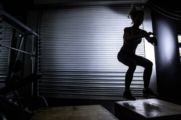 Mid adult female athlete in sportswear jumping onto plyo box holding kettlebell in warehouse gym