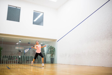 Fototapeta premium Female squash player swinging racket on court with glass back wall revealing gym and potted plant