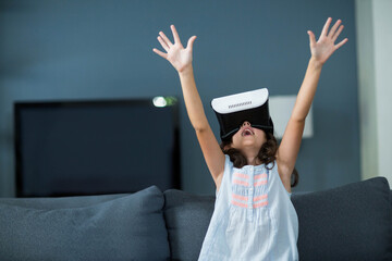 Female child raising arms exploring virtual reality headset on sofa in living room with TV screen