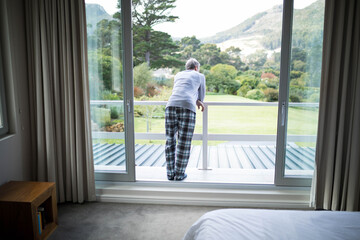 Senior man wearing pajamas leaning on balcony railing gazing at manicured lawn through glass doors