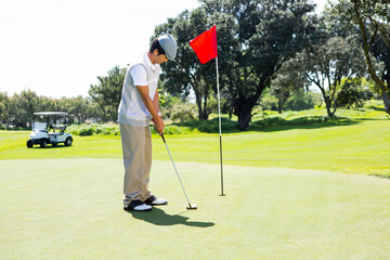Asian man aligning putter with golf ball on green by red flag with golf cart behind