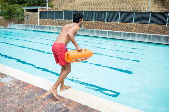 Mid-adult male lifeguard standing poolside holding rescue buoy by lanes in red trunks, copy space
