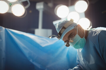 Male gowned surgeon in OR using loupe leaning over draped patient under lights, copy space