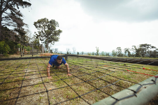 Male crawling under low net obstacle with red straps on grassy outdoor course, copy space