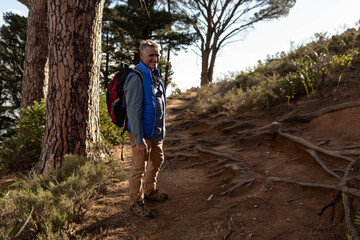 Mature male hiker standing on wooded trail wearing hiking boots, carrying red backpack, copy space