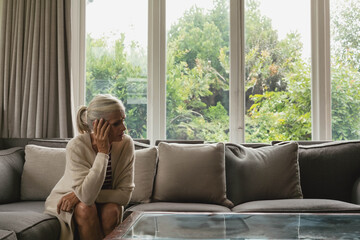 Senior woman sitting on sectional sofa at home leaning forward holding face near glass coffee table