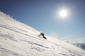 Snowboarder carving snowy mountainside under bright sun with shallow tracks near conifer tops