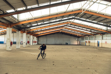 Man performing BMX bicycle stunt inside industrial warehouse wearing dark t-shirt and pants