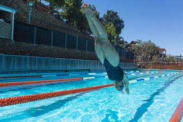 Female athlete wearing swimsuit, swim cap and goggles diving into outdoor pool lane with lane ropes